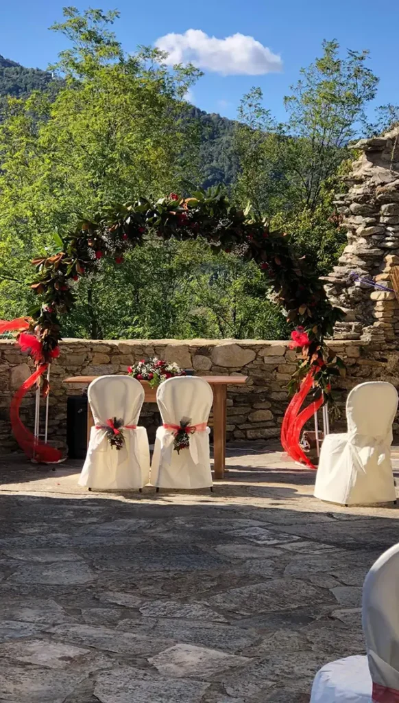 Arco decorativo con fogliame e nastri rossi nel prato davanti al Castello medievale con sedute bianche e muretto in pietra.