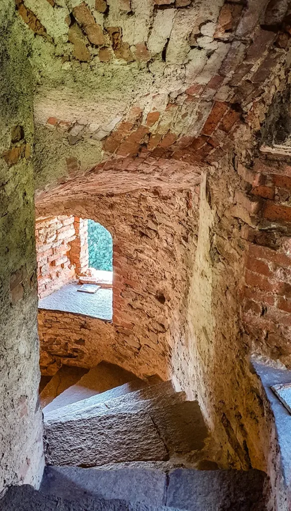 Interno del castello medievale con scala in pietra, muri in muratura antica e volta ad arco illuminata da una piccola apertura.