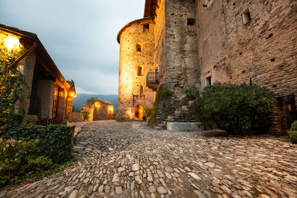 Cortile acciottolato del Castello medievale con torre cilindrica in pietra, portale ad arco illuminato, scala esterna e murature in pietra a vista al crepuscolo.