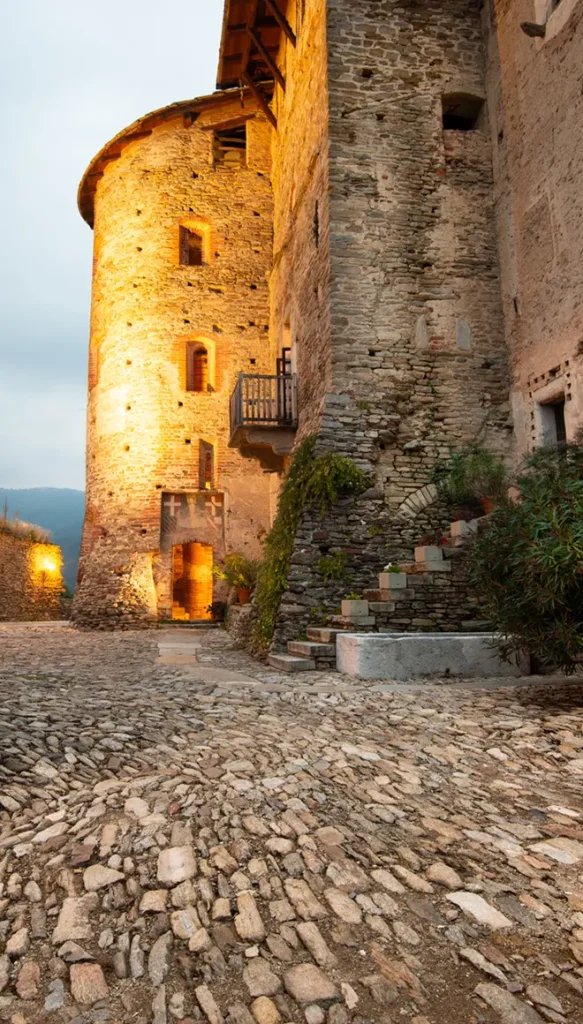 Cortile interno del Castello di Bagnolo Piemonte con torre medievale in pietra illuminata al crepuscolo
