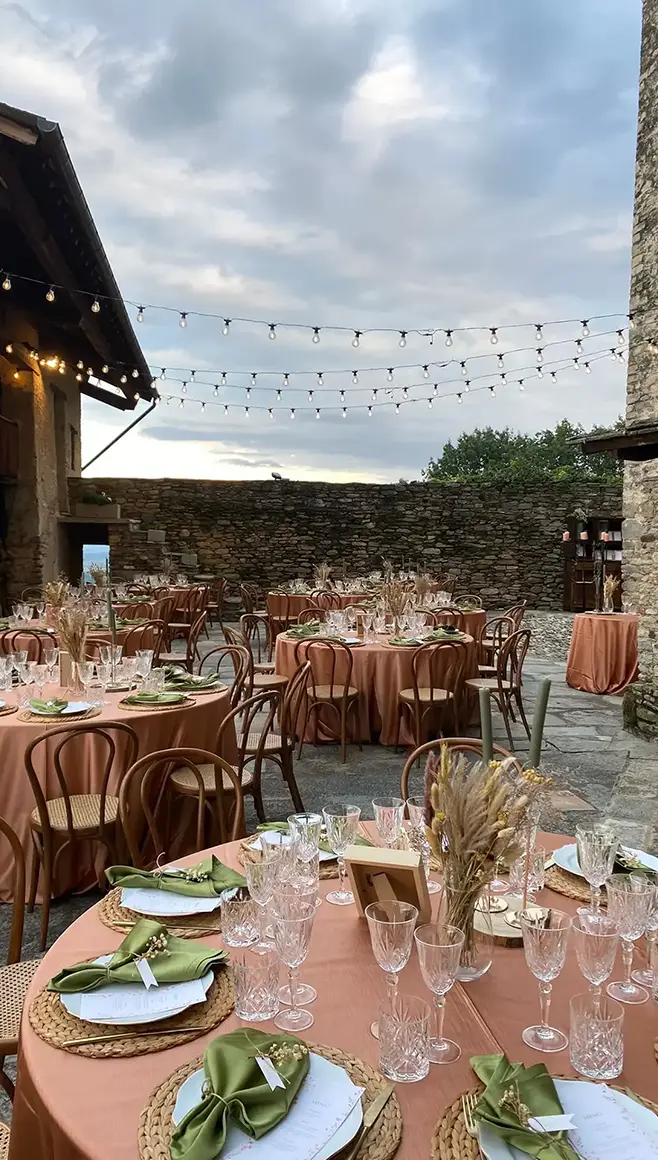 Tavoli rotondi apparecchiati nella corte interna del Castello medievale con murature in pietra e fili di luci sospese.