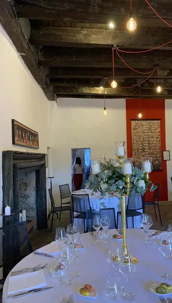 Sala del Castello medievale con soffitto a travi in legno, camino in pietra e tavoli rotondi apparecchiati con centrotavola floreali.