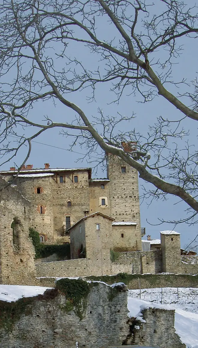 Veduta del castello medievale con torri e mura in pietra, parzialmente coperto dalla neve, osservato da distanza con alberi spogli in primo piano.