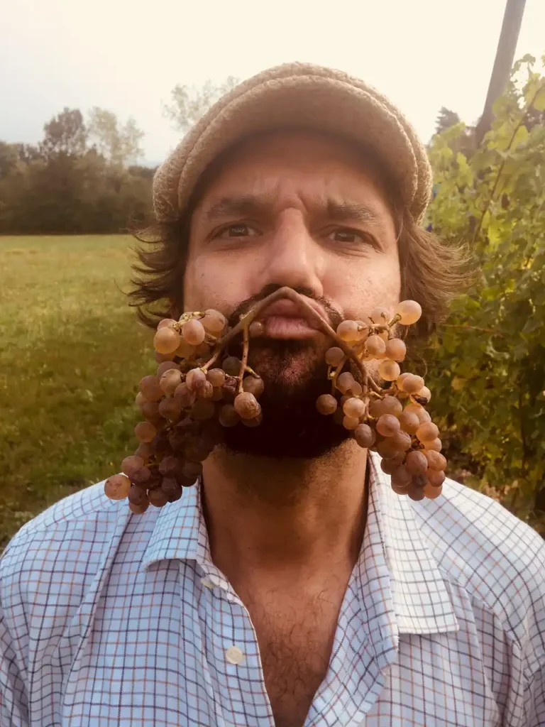 Fotografia di Hilario Isola realizzata tra i filari del vigneto. Lo scatto lo ritrae in un momento giocoso, con due grappoli d’uva appoggiati al volto come una barba improvvisata. L’immagine comunica una presenza libera, creativa e autentica, in sintonia con il paesaggio agricolo e con il carattere personale del suo lavoro tra arte e vino.